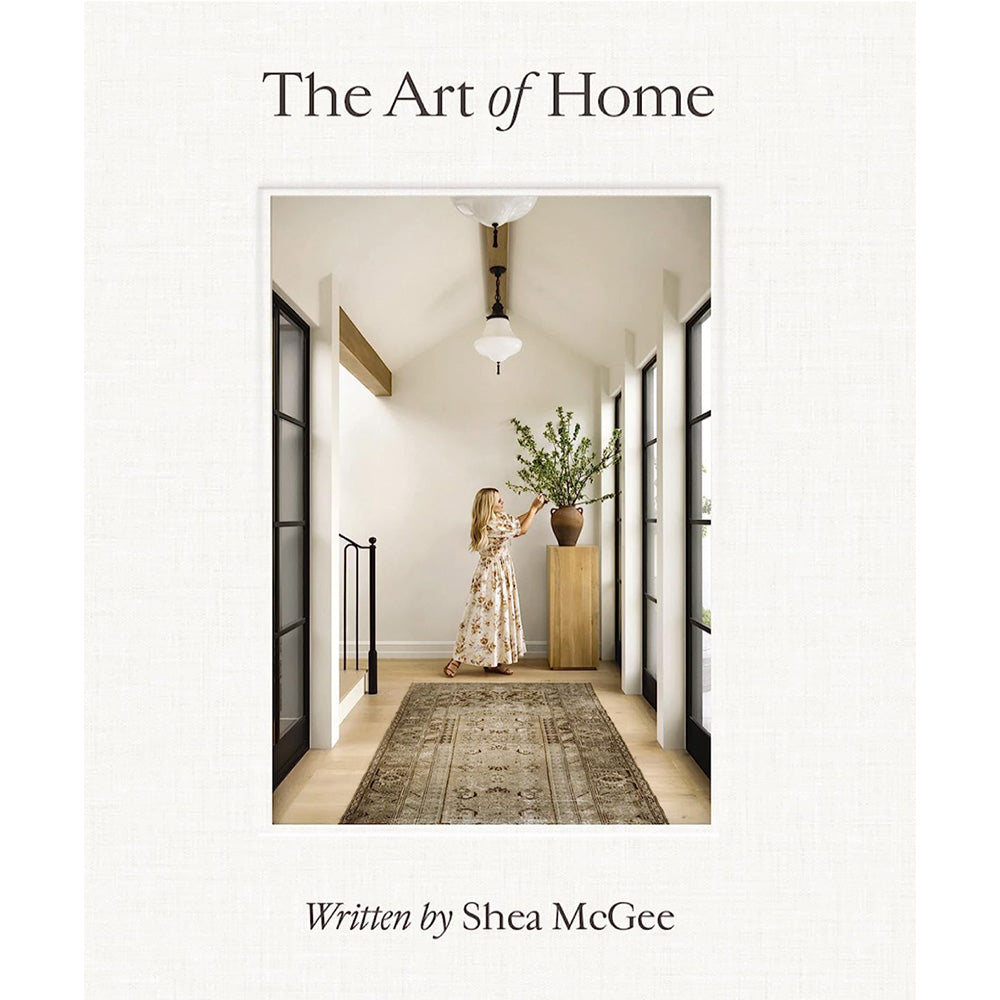 Photo of the cover of The Art of Home book written by Shea McGee showing a photo of Shea McGee fixing a vase of greenery which stands on a timber plinth.  There are black metal windows either side and a brown vintage floor runner on the timber floor.