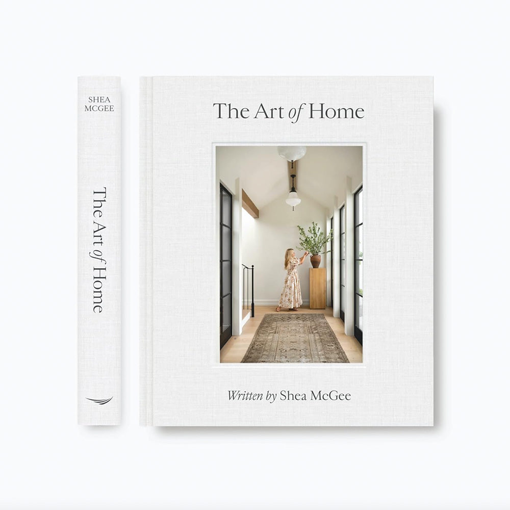 Photo of the side binding edge of The Art of Home book as well as a photo of the cover of The Art of Home book written by Shea McGee showing a photo of Shea McGee fixing a vase of greenery which stands on a timber plinth.  There are black metal windows either side and a brown vintage floor runner on the timber floor.