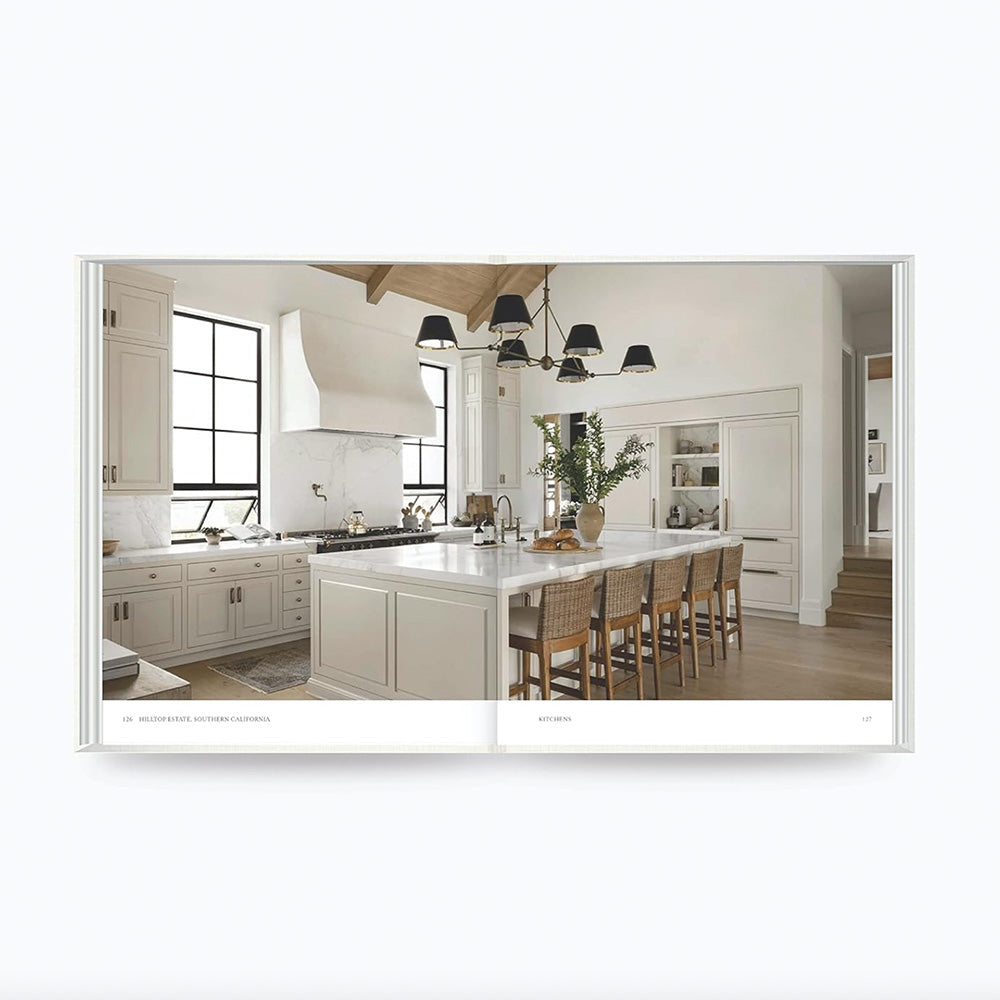 Photo of the inside pages of the Art of Home book showing a kitchen with white cabinets, natural timber bar stools, a black pendent light and timber floors.