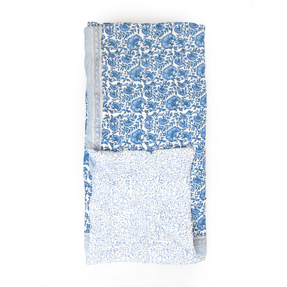 Photo of a blue and white floral quilted bedspread folded up with one corner turned upwards to show the reverse side of the quilt being a white background with soft blue floral print.
