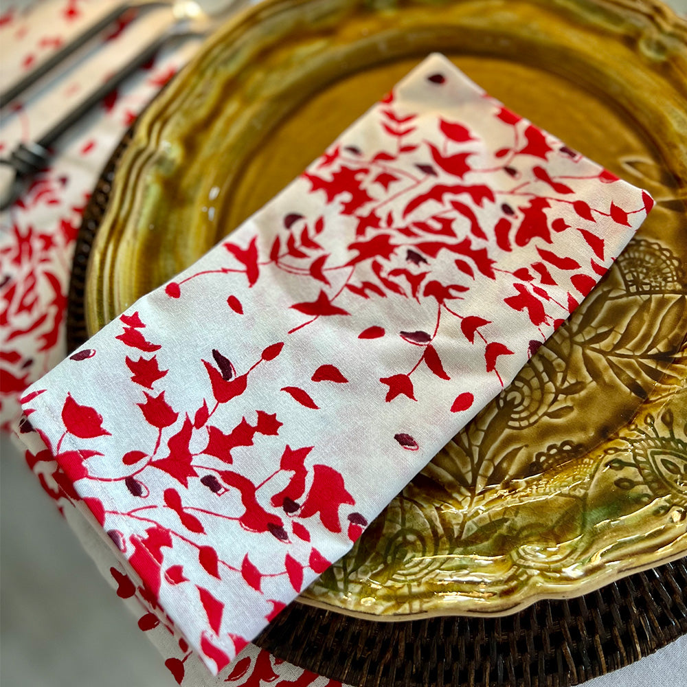 Close up photo of a folded white, red and deep burgundy red floral cotton napkin sitting on a spice coloured ceramic plate.