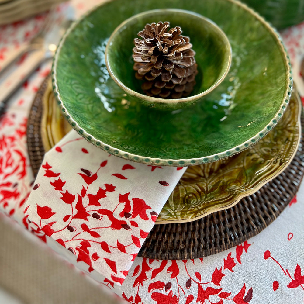 Photo of a folded white, red and deep burgundy red floral cotton napkin sitting under a green bowl on a Christmas table setting with the same fabric tablecloth on the table.