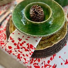 Photo of a folded white, red and deep burgundy red floral cotton napkin sitting under a green bowl on a Christmas table setting with the same fabric tablecloth on the table.