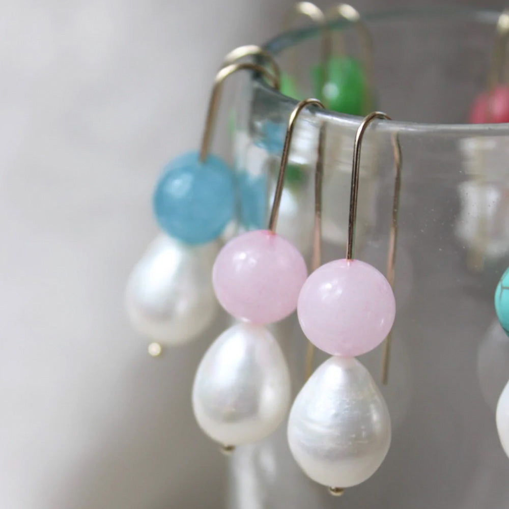 Photo of gold hook earrings with a large teardrop shaped freshwater pearl and a round pale pink coloured agate stone.  The earrings are hanging over the side of a glass and you can see the other colour options of blue, green and deep pink in the background. 