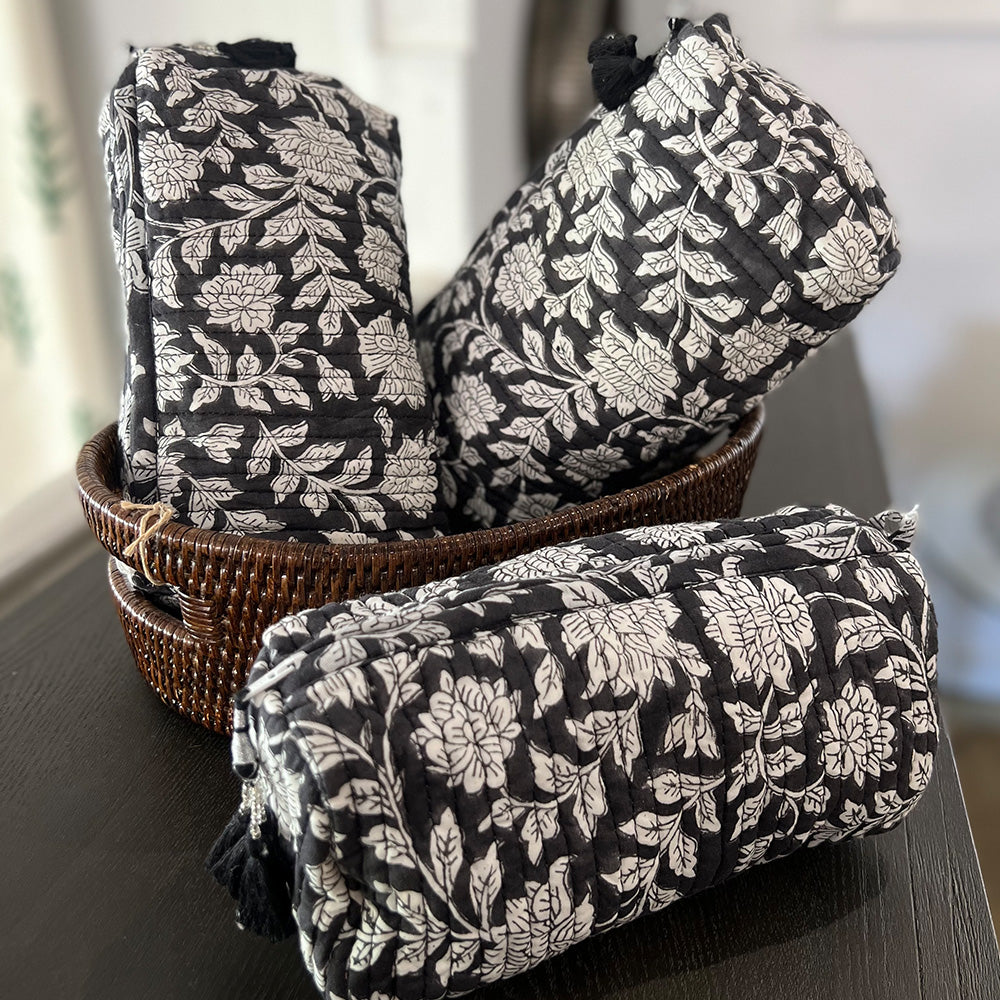Photo of three blue charcoal floral quilted cosmetic pouches showing the three different sizes.  Two pouches are inside a dark rattan basket and the third is sitting on a dark timber bench.