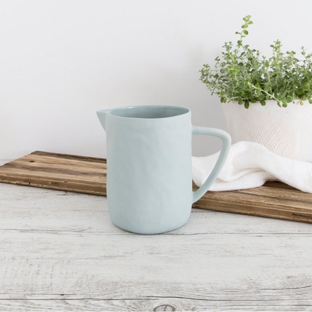 Small duck egg blue ceramic milk jug with a matt finish on outside of jug and shiny ceramic gloss finish on the inside.  The jug is on a whitewash timber bench beside a timber chopping board, white napkin and white vase with greenery. 
