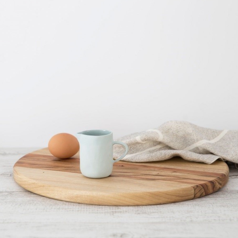 Small duck egg blue ceramic milk jug with a matt finish on outside of jug and shiny ceramic gloss finish on the inside.  The jug is on a whitewash timber bench on top of a round timber chopping board, beige teatowel and one egg.
