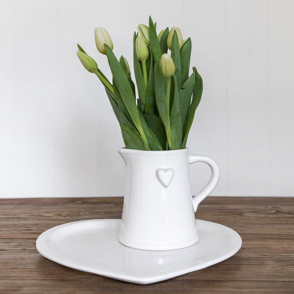 Photo of a white ceramic jug with raised heart detail on the side of the jug, and there are white tulip flowers in the jug.