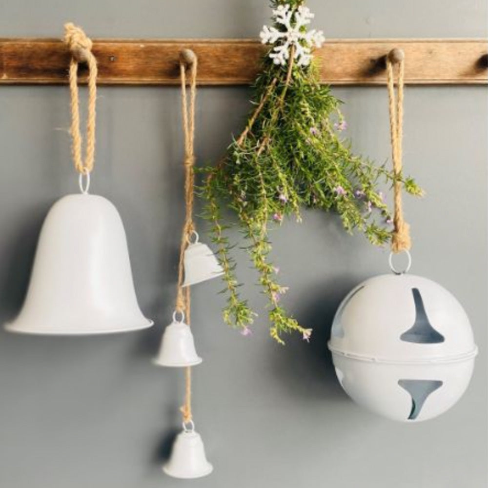 Photo of white painted metal Christmas hanging decorations of large bell, three small bell garlands and a large round ball with cutouts, all hanging from a timber peg rail with string.  There is also a bunch of heather plant hanging on the peg rail with a white paper snowflake