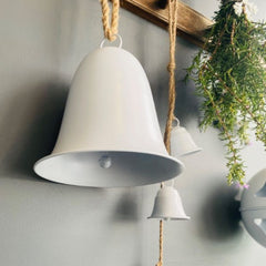 Close up photo of white painted metal Christmas hanging decorations of large bell, three small bell garlands and a large round ball with cutouts, all hanging from a timber peg rail with string.  There is also a bunch of heather plant hanging on the peg rail with a white paper snowflake.