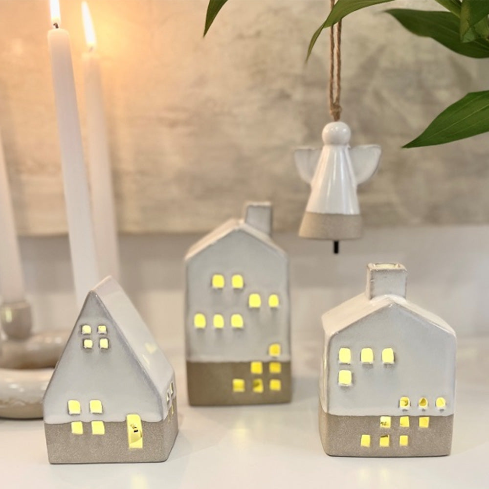 Photo of four ceramic Christmas decorations to show the featured item the Woodstock A Frame House.  Three separate shaped houses with cutouts for windows showing the battery operated light shining through the windows, as well as a cream ceramic hanging angel bell decoration.