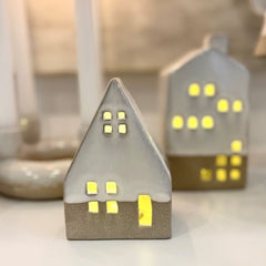 Close up of white ceramic glazed A-frame house with battery operated light shining through the window and door cut outs.  The tall ceramic house sits in the distance.