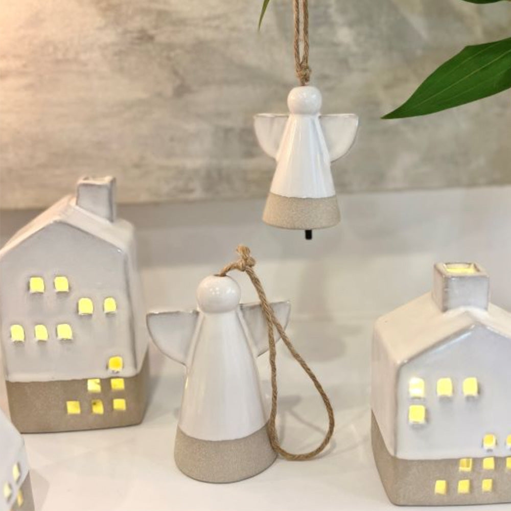Photo of white ceramic angel hanging ornament sitting on the table as well as the angel ceramic ornament hanging behind. There is a tall ceramic house sitting to the left and a small ceramic house sitting to the right.