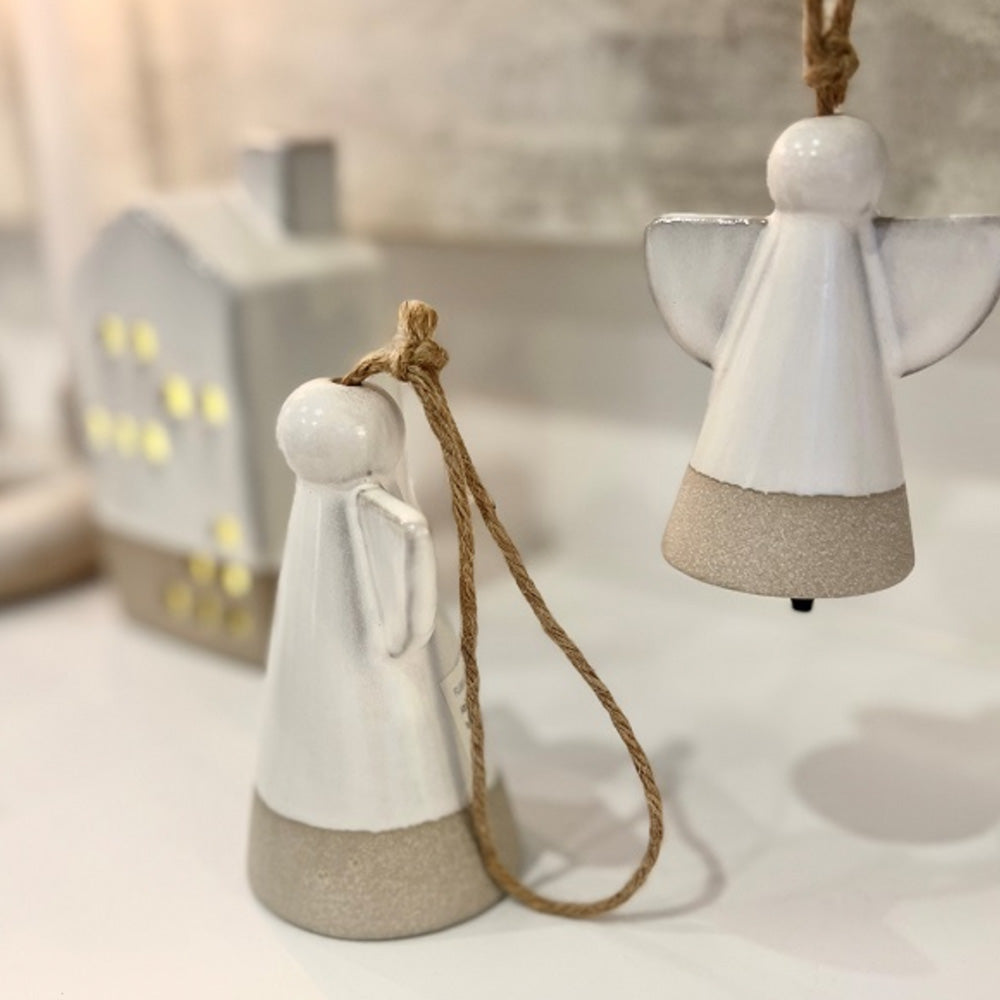 Close up photo of white ceramic angel hanging ornament sitting on the table as well as another angel ceramic ornament hanging behind. In the distance there is tall ceramic house sitting to the left.