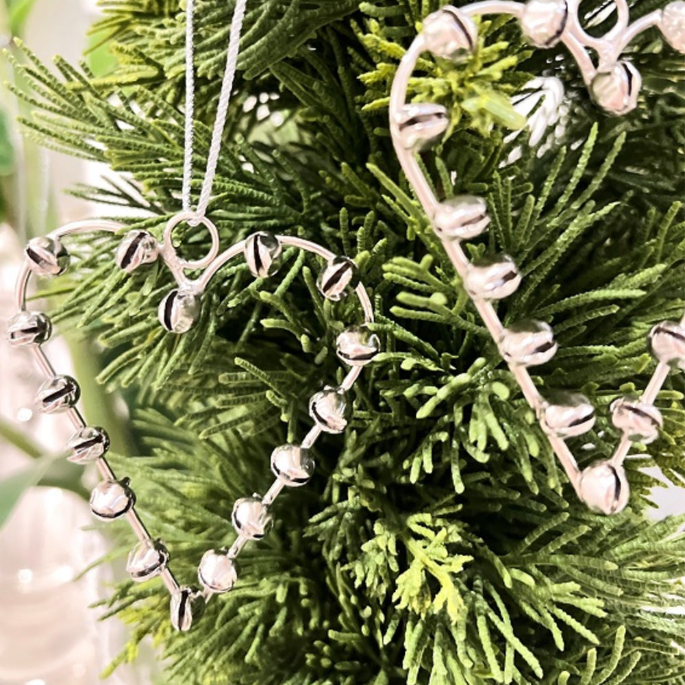Silver metal heart hanging Christmas decoration with small silver bells attached to the trim of the heart, hanging by string on a green Christmas tree.