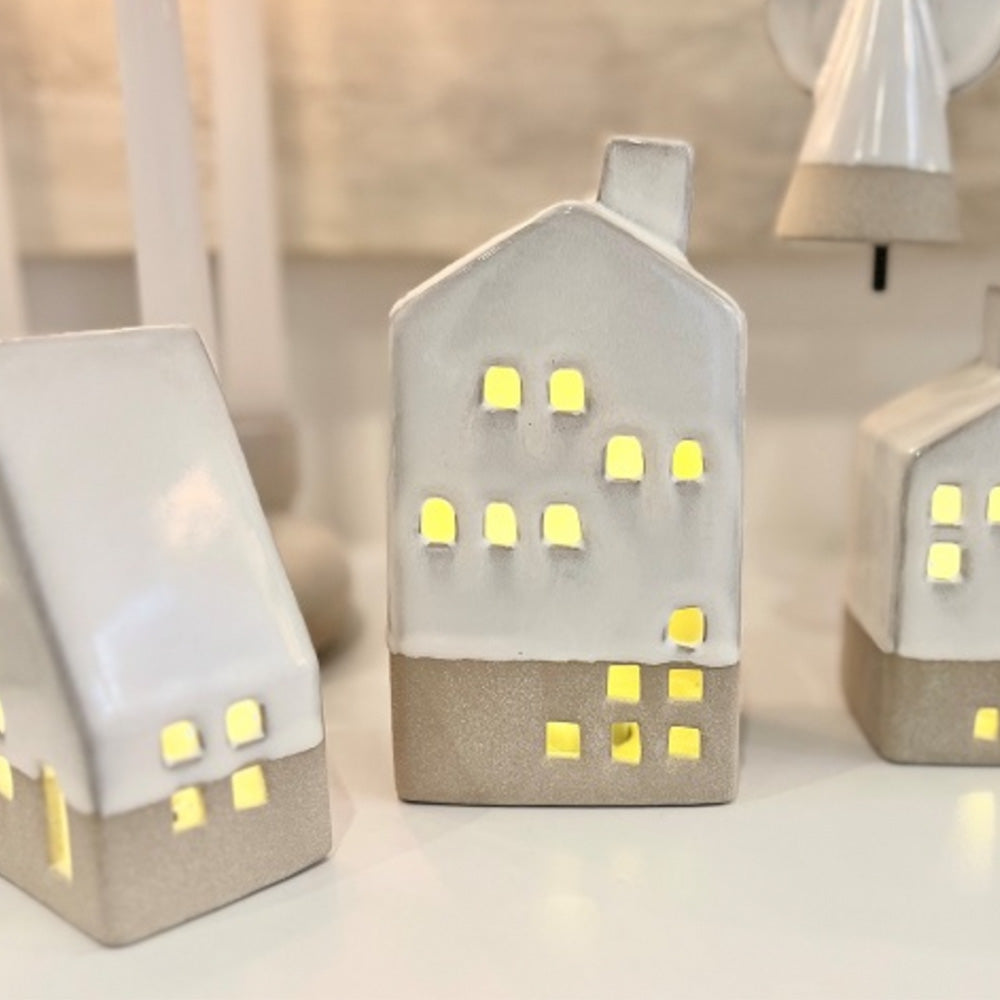 Close up of white ceramic tall house with battery operated light shining through the window and door cut outs.  The tall ceramic house is in the centre of the photograph with the A-frame house to the left and small house to the right as well as the angel ceramic ornament hanging behind.