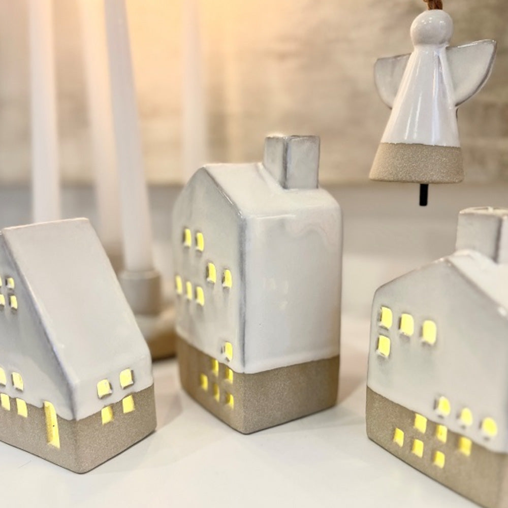 Close up of white ceramic tall house with battery operated light shining through the window and door cut outs.  The tall ceramic house is in the centre of the photograph with the A-frame house to the left and small house to the right as well as the angel ceramic ornament hanging behind.