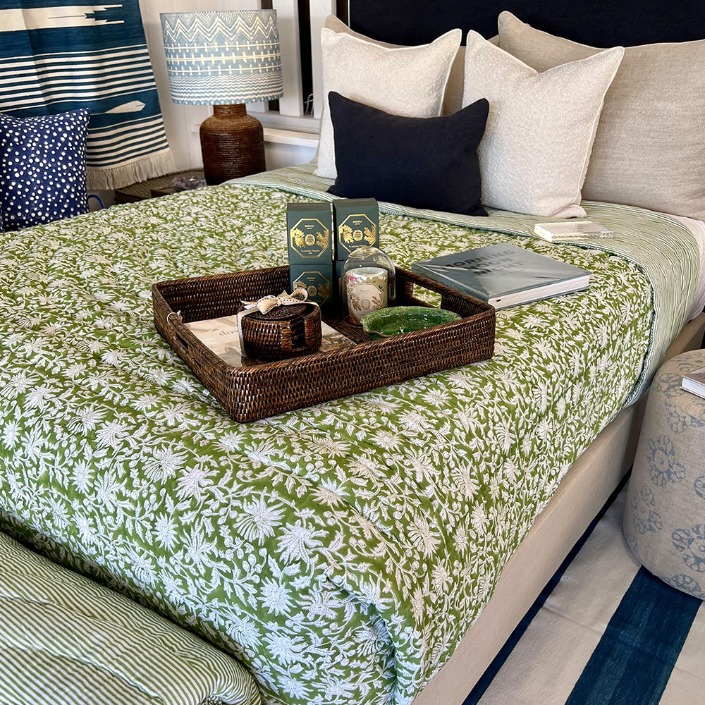 Photo of a forest green and white floral quilted bedspread over a bed.  There is a dark brown rattan tray on the bed with items in it such as dark rattan coaster set, green bowl and candles.