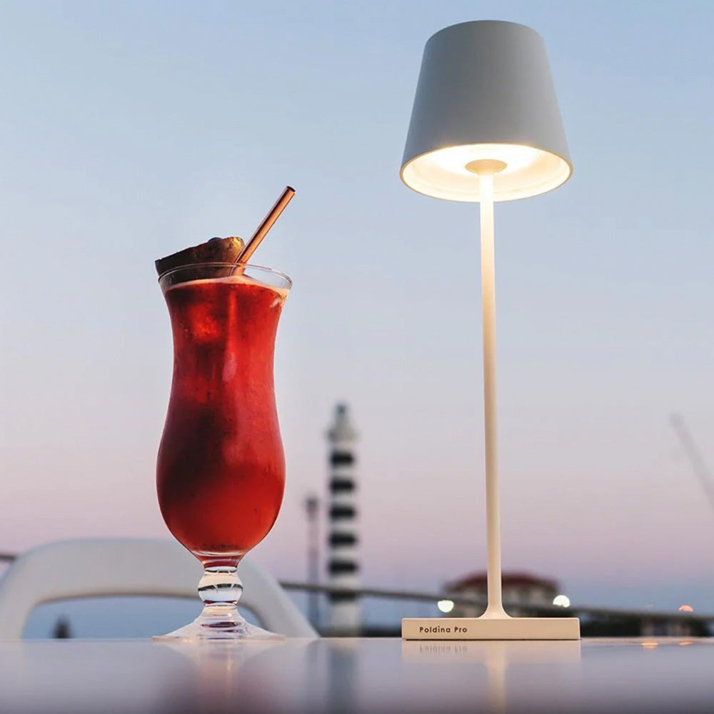 Photo of a white metal battery operated table lamp sitting on a bench and there is a red coloured cocktail drink in a tall glass sitting beside it.