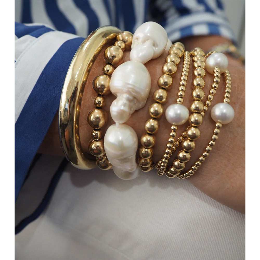 Photo of a woman's arm showing a collection of gold and pearl bracelets and bangle.  The gold ball chain bracelet is featured in the middle of this collection.