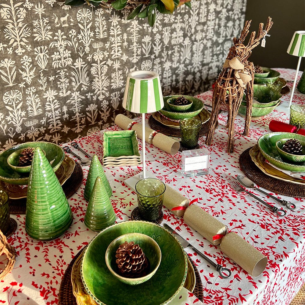 Photo of a Christmas table setting with a cotton tablecloth and napkins in white, red and deep burgundy red floral fabric.  On the table are green bowls, dark rattan placemats, two green and white battery operated table lights and two twig reindeer table ornaments.