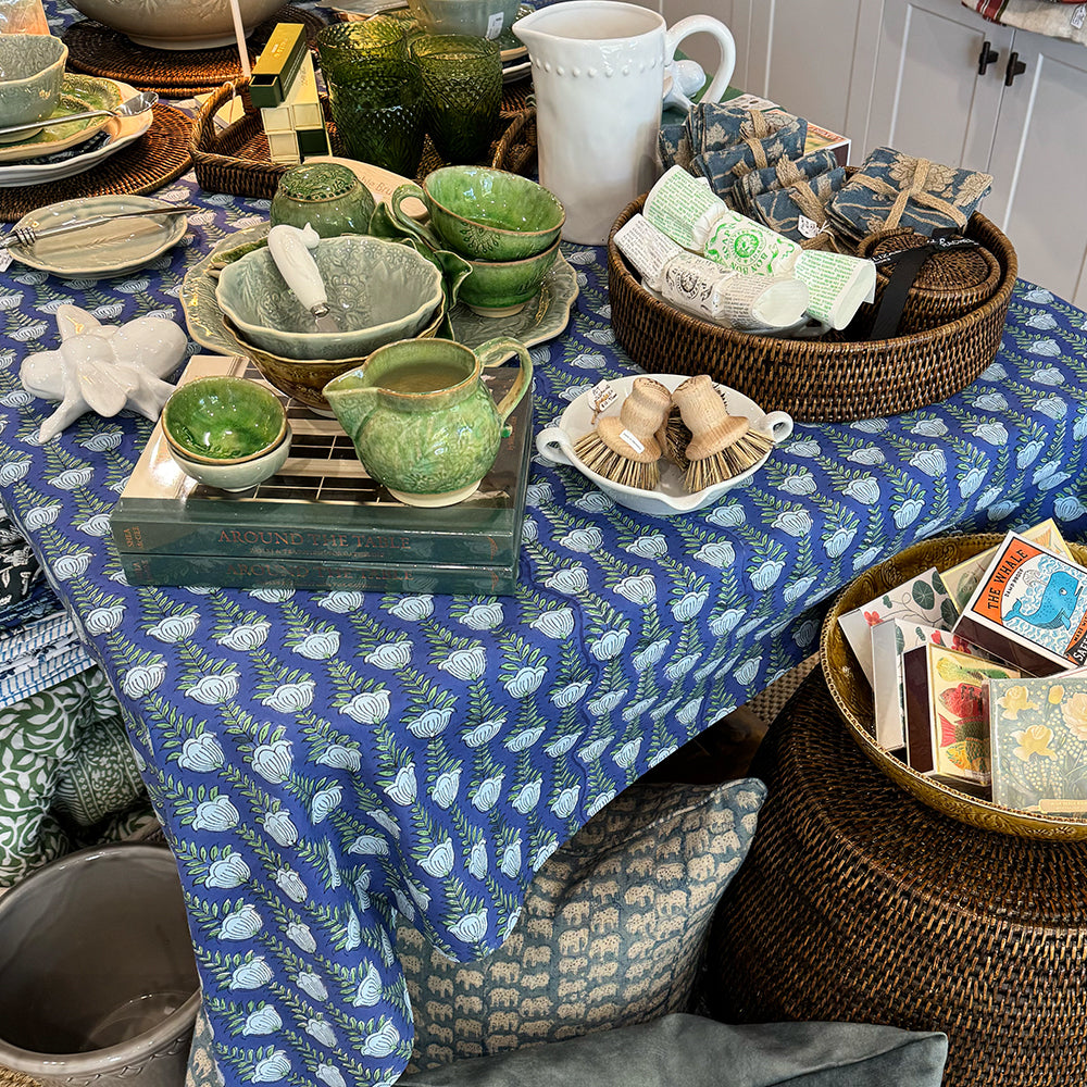 A styled tabletop display featuring a blue and white patterned tablecloth, green ceramic dishes, woven baskets, and home decor accessories in a rustic shop setting.