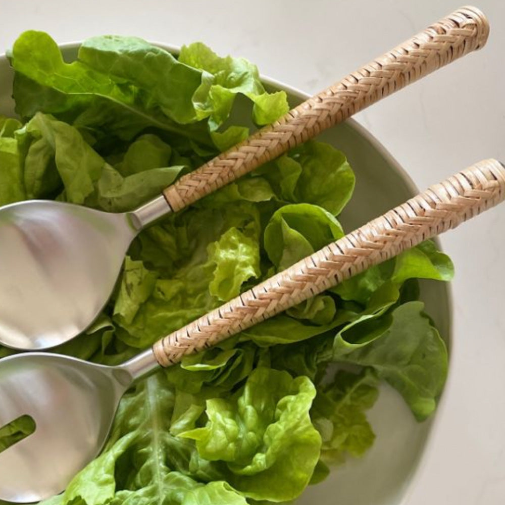 Brushed silver salad servers with natural rattan woven around the handles sitting in a white salad bowl on top of green lettuce leaves.
