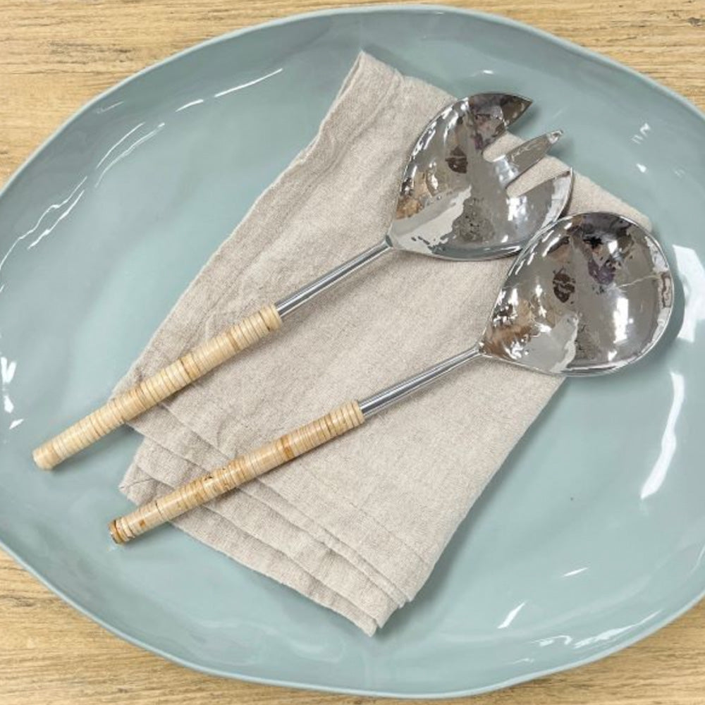 Shiny stainless steel salad servers with a beaten detail finish and handles are wrapped in natural rattan.  The servers are sitting on a pale blue shiny ceramic platter and beige linen napkin.