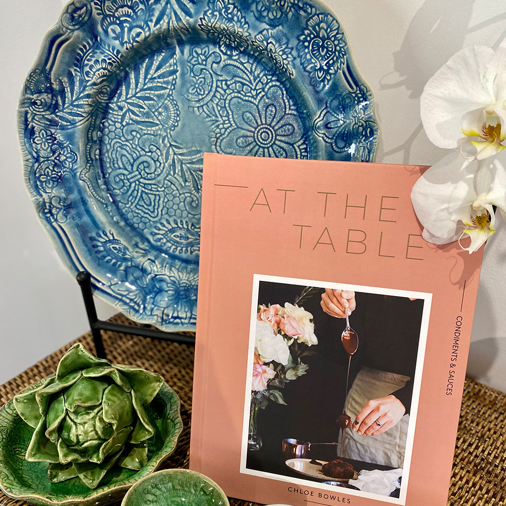 Photo of front cover of At The Table recipe book sitting beside a blue plate and behind some green ceramic bowls